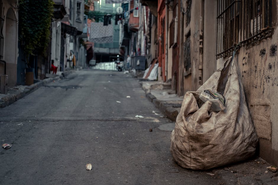 A concentrated scene of overflowing waste bins and scattered rubbish on a paved street corner in front of a commercial building. The central large grey bin is filled with mixed paper and cardboard, wi
