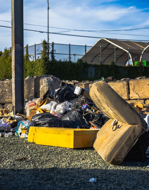 A pile of mixed waste materials and packaging, including cardboard boxes, paper bags, and plastic wrapping, accumulated at the base of a brick wall in an outdoor setting. The cardboard boxes vary in s