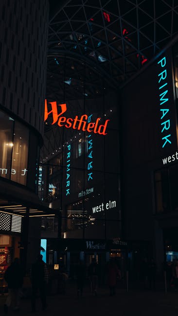 Inside a modern commercial building with a high glass ceiling and dark structural elements, illuminated by neon signs displaying the words 'Westfield', 'Primark', 'West Elm', and 'Primark' in vivid bl