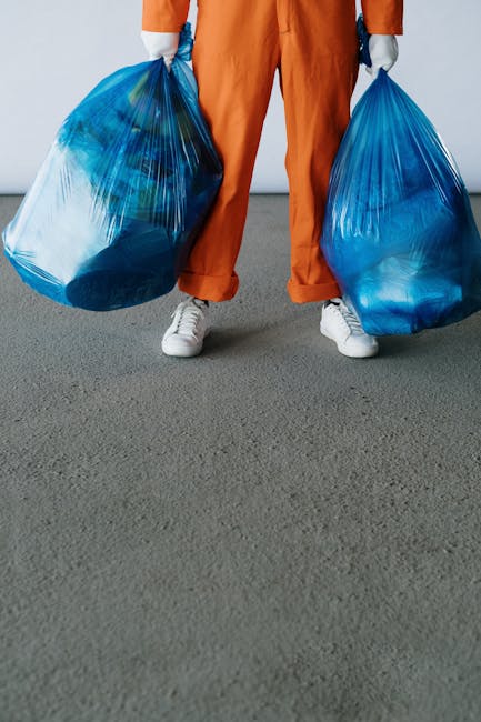 A person dressed in orange work overalls and white sneakers is standing on a grey outdoor surface, holding two large blue plastic trash bags filled with waste, one in each hand. The bags are semi-tran