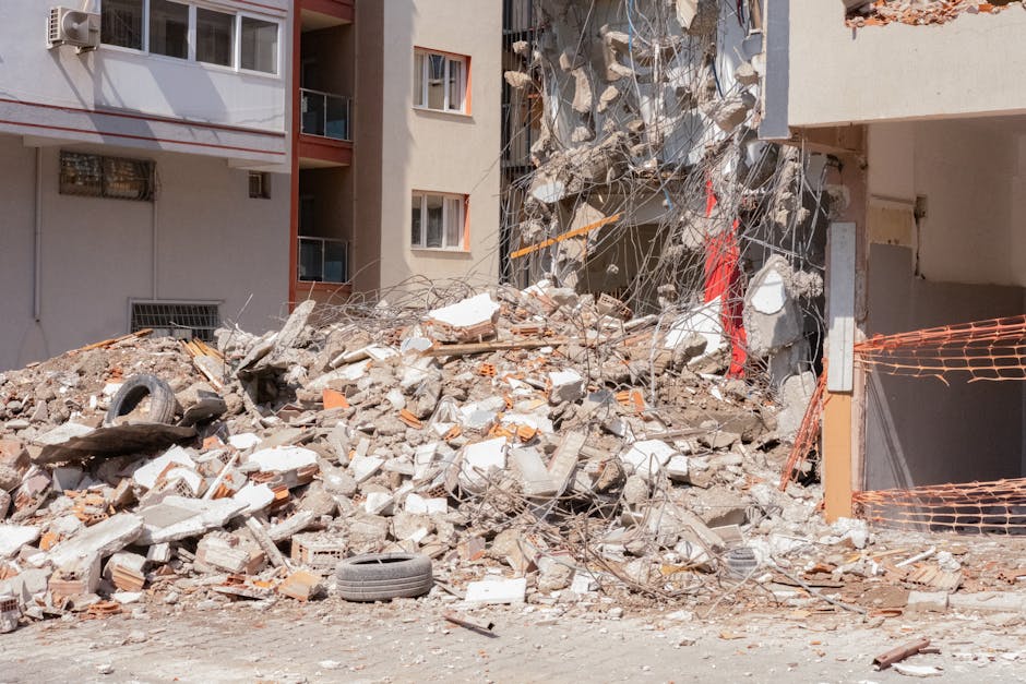 A pile of rubble and debris, consisting of broken bricks, concrete fragments, and assorted construction materials, is scattered across the ground in front of a partially damaged multi-storey residenti